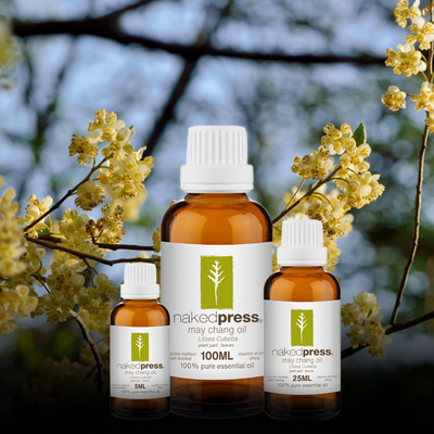 Three bottles of NakedPress May Chang Essential Oil with a label showing a plant and the text 'May Chang Oil'. The background has a blurred image of flowers.